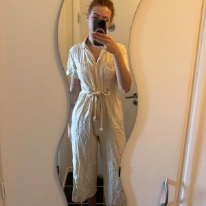 Beige jumpsuit från Make Way - Säljer en beige jumpsuit från Make Way med kort ärm, krage och knappar framtill. Jumpsuiten har knytskärp i midjan och raka ben i croppad längd. Perfekt för en avslappnad och stilren look.
