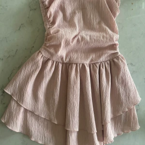 Ljusrosa volangklänning med smock - Supersöt ljusrosa kort armlös klänning med smock i ryggen och dubbla volanger i kjolen. Endast använd 1-3 gånger (minns inte riktigt).