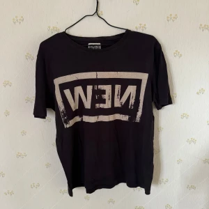 Svart t-shirt från Weekday - Svart t-shirt från Weekday med stort beige tryck på framsidan där det står 'NEW' spegelvänt. Klassisk rund hals och korta ärmar. Perfekt för en avslappnad och cool stil.