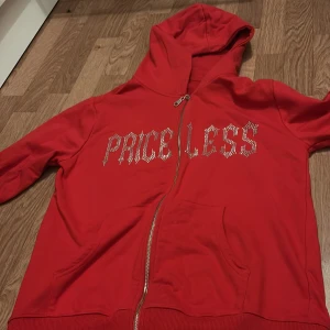Röd hoodie med texten 'PRICELESS' - Röd hoodie med huva och dragkedja framtill. Texten 'PRICELESS' i glittriga stenar på bröstet. Mjuk bomullskänsla och två fickor framtill. Perfekt för en avslappnad stil.