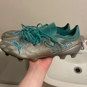 Limited edition Puma Ultra Collab med Mercedesfotbollsskor i silver och turkos - Säljer ett par Puma Ultra fotbollsskor i silver med turkosa detaljer och snörning. Skorna har rund tå, platt sula med dobbar och är designade för maximal fart på planen. Perfekta för dig som vill sticka ut med stil och prestanda.