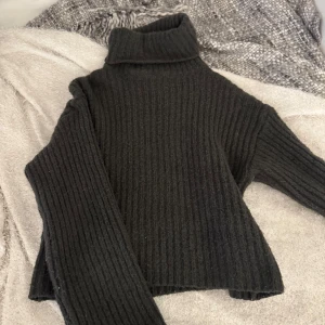 Mörkgrå  stickad polotröja - Mysig svart stickad polotröja med bred ribbad struktur och hög krage. Tröjan har långa ärmar och en något boxig, croppad passform. Perfekt för kyliga dagar och enkel att styla till olika outfits.