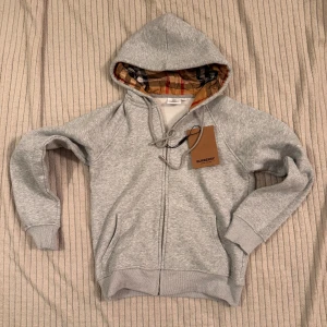 Burberry med dragkedja - Grå zip hoodie från Burberry i storlek L, men den är liten i modellen och passar bättre som S. Helt ny, endast testad – säljes då den tyvärr är för liten för mig. Riktigt snygg och stilren med exklusiv känsla.