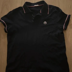 Svart pikétröja från Moncler - Säljer en svart pikétröja från Moncler med klassisk krage och knappar framtill. Röd och vit randig detalj på krage och ärmslut. Perfekt för en stilren look.