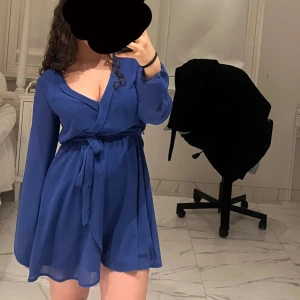 Blå omlottklänning med bälte - NYSKICK! Säljer en blå omlottklänning med knytband i midjan och långa ärmar. Klänningen har en v-ringad hals och är tillverkad i ett lätt, flödande material som ger en luftig känsla. Funkar från Xs till M