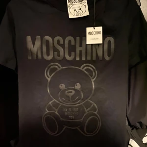 Svart t-shirt med björntryck från Moschino - Cool svart t-shirt från Moschino med stort björntryck och logga framtill. T-shirten har klassisk passform och är tillverkad i mjuk bomull. Perfekt för dig som vill sticka ut med en ikonisk design. Varan är äkta, tveka inte att kontakta vid flera funderingar. Orginalpris 2999kr. Helt oanvänd. Storlek 48 vilket motsvarar M