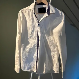 Vit skjorta med blå detaljer från Savvy Citizen - Vit skjorta från Savvy Citizen i storlek medium. Skjortan har lång ärm och blå kontrastdetaljer längs knappslå och krage. Klassisk krage och knäppning framtill, perfekt för en stilren look.