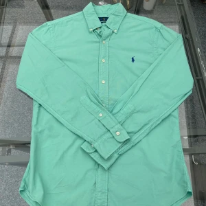 Grön skjorta från Ralph Lauren - (ÖPPEN FÖR BUD)Snygg grön skjorta från Ralph Lauren i slim fit-modell. Skjortan har lång ärm, klassisk krage och knappar framtill. Broderad blå logga på bröstet ger en stilren touch. Perfekt för dig som gillar klassisk och stilren design.  