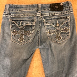 Ljusblå jeansbyxor med broderade bakfickor från Miss Me - Säljer ett par lowrise ljusblå jeansbyxor från Miss Me med raka ben och snygga broderade detaljer på bakfickorna. Fickorna har korsmönster med nitar och stenar för extra bling. Klassisk femficksmodell och straight passform.