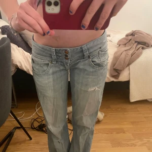Lågmidjade ljusblå jeans med slitningar - Säljer ett par lågmidjade ljusblå jeans med raka ben och slitna detaljer framtill. Jeansen har  femficksdesign, trippla knappar i midjan och en baggy passform. 