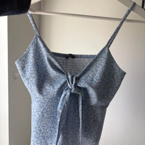 Blå blommig linnetopp med knyt - Söt blå linnetopp med små vita blommor och smala axelband. Toppen har knyt framtill och smockad rygg för extra komfort. Perfekt för varma dagar och enkel att matcha med jeans eller kjol.