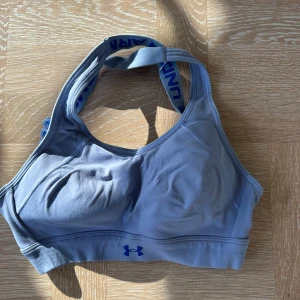 Blå/lila sporttopp från Under Armour - Sportig grå linnetopp från Under Armour med blå logga framtill och breda axelband med blå text. Perfekt för träning och har en tight passform som ger bra stöd. Snygg och enkel design som funkar till alla träningspass.