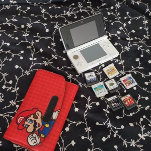 Vitt Nintendo DS 3S med spel och laddare. Röd Mario väska för spel. Fungerar utan anmärkning.  - Säljer en röd handväska med coolt Super Mario-motiv, perfekt för att förvara Nintendo DS/3DS och spel. Väskan är rektangulär med svart och blå detaljer, och stängs med kardborre. Materialet är syntet och ytan har ett diskret mönster. Passar dig som vill ha gamingprylar med stil.