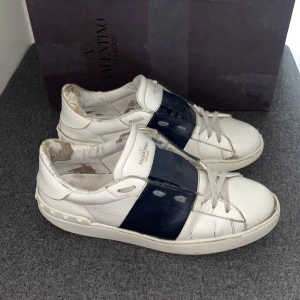 Valentino skor - Snygga Valentino Garavani Open sneakers i vitt skinn med bred marinblå rem över mitten. Klassisk rund tå, platt sula och coola nitar baktill. Skorna har snörning och en stilren design som sticker ut.