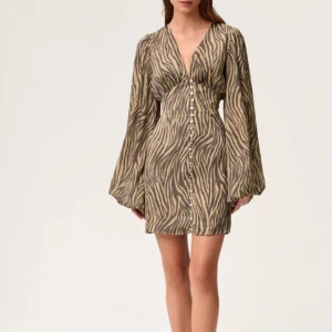 Zebramönstrad kortklänning med v-ringning - Supersnygg kort klänning med zebramönster i beige och svart. Djup v-ringning, långa vida ärmar och knappar framtill hela vägen ner. Storlek 32 som aldrig är andvänd och har prislappen kvar