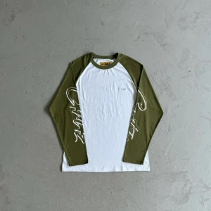 Corteiz långärmad tröja i vitt och olivgrönt - Snygg långärmad tröja från Corteiz med vita och olivgröna partier. Ärmarna har coolt grafittiliknande tryck och raglanmodell. Perfekt för dig som gillar streetwear och vill sticka ut lite extra. Mjuk bomullskvalitet och relaxed fit.