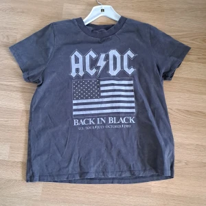 AC/DC Back in Black t-shirt - Grym svart AC/DC t-shirt med tryck från Back in Black-turnén 1980 och amerikansk flagga på bröstet. Klassisk rund hals och korta ärmar, perfekt för dig som gillar rock och vintage-stil.