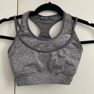 Grå sporttopp från Gymshark - Säljer en grå sporttopp från Gymshark med brottarrygg och sömlös design. Toppen är i stretchigt material och har en melerad look. Perfekt för träning och ger bra stöd. Snygg och enkel att matcha med träningskläder.