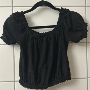 Svart blus med puffärm och broderi - Supersöt svart blus med puffiga korta ärmar och broderade detaljer. Modellen är croppad med resår i både halsringning och nederkant för en snygg passform. Perfekt för dig som gillar romantiska vibes och vill sticka ut lite extra.