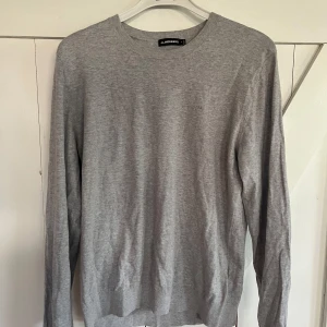 Grå långärmad tröja från J.Lindeberg - Snygg grå långärmad tröja från J.Lindeberg med diskret logga på bröstet. Tröjan har rund halsringning och är tillverkad i mjuk bomull, perfekt för en clean och stilren look. Passar dig som gillar enkel och klassisk stil. Passar också på golfbanan.