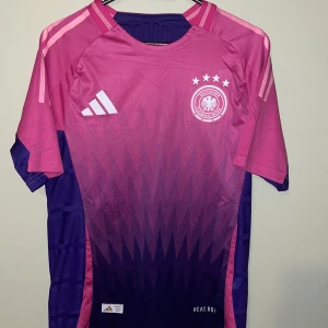 Tysklands fotbollströja Adidas rosa/lila - Säljer en Tyskland fotbollströja från Adidas i rosa och lila med gradientmönster. Tröjan har korta ärmar, klassiska Adidas-ränder på axlarna och Tysklands landslagsemblem med fyra stjärnor på bröstet. Tillverkad i lätt och ventilerande funktionsmaterial.