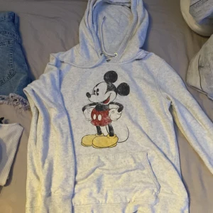 Grå hoodie med Mickey Mouse-tryck - Supermysig ljusgrå hoodie från Disney med stort tryck av klassiska Mickey Mouse på framsidan. Tröjan har huva och känguruficka, perfekt för chill dagar. Mjuk bomullskänsla och långärmad modell. Passar dig som gillar retro och tecknade motiv!