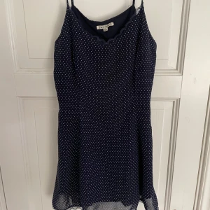 Mörkblå prickig klänning Lucca Couture - Supersöt mörkblå kortklänning från Lucca Couture med vita prickar, tunna axelband och spetsdetaljer vid halsringning och fåll. Klänningen har en lätt och luftig känsla, perfekt för varma dagar. Prickmönstret ger en lekfull vibe och spetsen adderar en romantisk touch.