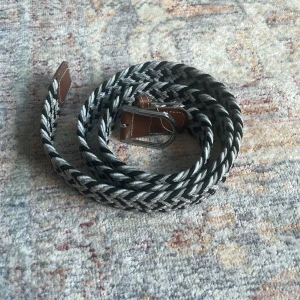 Svart och grått flätat bälte - Säljer ett snyggt flätat bälte i svart och grått med silverfärgat spänne och bruna skinn-detaljer vid ändarna. Perfekt accessoar för att lyfta din outfit med en cool och modern touch.