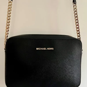 Svart axelväska från Michael Kors - Stilren svart axelväska från Michael Kors i skinn med guldfärgade kedjedetaljer på axelremmen. Väskan har en enkel, fyrkantig form och diskret logga framtill. Perfekt storlek för det viktigaste och en klassisk look som funkar till allt.