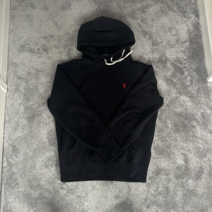 Svart hoodie från Polo Ralph Lauren - Svart hoodie från Polo Ralph Lauren med klassisk röd logga broderad på bröstet🐎 Tröjan har vit snörning i huvan och känguruficka. Perfekt för en chill och stilren look✅