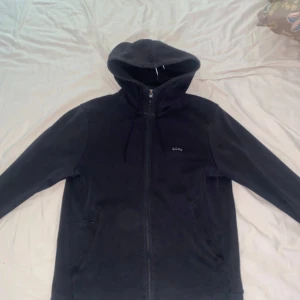 Svart hoodie från BOSS med dragkedja - Svart hoodie från BOSS med hel dragkedja, huva med snörning och diskret BOSS-logga på bröstet. Tröjan har långa ärmar och två fickor framtill. Insidan av kragen har en grön detalj. Perfekt för en chill och stilren look.