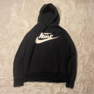 Svart Nike hoodie  - Svart hoodie från Nike i storlek XS med stor vit Nike-logga och swoosh på bröstet. Klassisk känguruficka framtill, huva med snörning och långa ärmar. Perfekt för dig som gillar sportig och avslappnad stil.