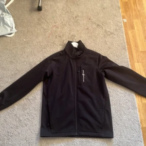 Svart vindjacka från Sail Racing M - Svart vindjacka från Sail Racing i storlek medium. Jackan har hög krage, dragkedja framtill och en bröstficka med dragkedja. Materialet är mjukt och smidigt, perfekt för blåsiga dagar. Snygg och stilren design med diskret logga på bröstet.