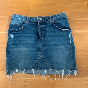 Blå jeanskjol med fransig kant - Snygg blå jeanskjol med rå, fransig nederkant och slitna detaljer. Klassisk femficksmodell med bälteshällor och dragkedja fram. Perfekt för dig som gillar en avslappnad och trendig stil. Funkar både som lågmidjad och högmidjad beroende på vad man föredrar💕 Säljer då den var för stor 