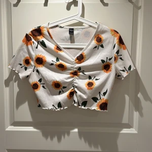 Vit croppad topp med solrosor  - Superfin croppad topp med solrosmönster och v-ringning. Toppen har korta ärmar, rynkdetalj framtill och är gjord i mjuk, ribbad bomull. Perfekt för dig som vill sticka ut med färgglada blommor och somrig vibe!