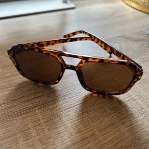 Leopardmönstrade solglasögon retro - Snygga solglasögon med leopardmönstrad båge i brunt och svart. Modellen har en cool retroinspirerad form med markerad brygga och bruna glas. Perfekt accessoar för att lyfta din stil till nästa nivå.