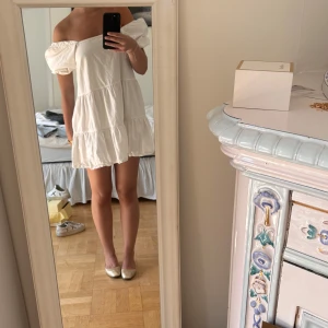 Vit offshoulder kortklänning - Supersöt vit kortklänning med offshoulder och puffiga ärmar. Klänningen har volangdetaljer i flera lager och en luftig passform som känns riktigt somrig. Perfekt för dig som gillar en romantisk och trendig stil men även perfekt inför studenten 