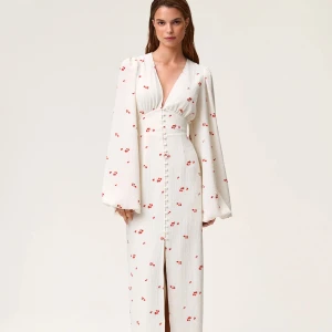 Vit långklänning med blommigt mönster - Superfin vit långklänning med små röda blommor, djup v-ringning och markerad midja. Klänningen har långa puffärmar och knappar framtill hela vägen ner, samt en slits. Perfekt för sommarens alla tillfällen! Använd vid ett tillfälle så fortfarande nytt skick. 