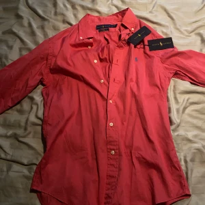 Röd skjorta från Ralph Lauren Slim Fit - Riktigt snygg röd skjorta från Ralph Lauren i slim fit-modell. Klassisk krage, långa ärmar och knappar framtill. Broderad blå logga på bröstet och tillverkad i lätt bomullstyg. Perfekt för dig som gillar stilrena och tidlösa plagg.