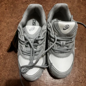 Asics gel NYC - Snygga sneakers från Asics i grått och vitt med meshpaneler och detaljer i syntet och mocka. Skorna har snörning, rund tå och en chunky vit sula som ger en sportig vibe. Perfekta för dig som gillar stilrena och bekväma sneakers till vardags.