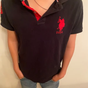 Svart US Polo Assn pikétröja - Svart pikétröja från US Polo Assn med röd broderad logga och texten USPA på bröstet. Kragen har röda detaljer och knappar framtill. Klassisk passform och korta ärmar, perfekt till jeans eller shorts.