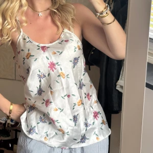 Blommig vit linnetopp med tunna axelband - Supersöt vit linnetopp med tunna axelband och blommigt mönster i lila, rosa, gult och grönt. Toppen har en lös passform och är perfekt för varma dagar. Materialet känns lätt och svalt mot huden.