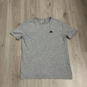 Grå t-shirt från Kappa - Snygg grå t-shirt från Kappa med klassisk logga broderad på bröstet. T-shirten har rund halsringning och korta ärmar. Perfekt basic-plagg med stilren look och mjukt material.
