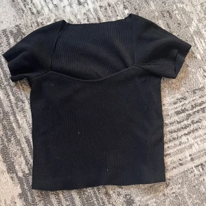 Svart ribbad topp med kort ärm - En svart, ribbad topp med korta ärmar och markerad byst. Snygg, figurnära passform och fyrkantig halsringning. Perfekt att styla med jeans eller kjol för en trendig look.