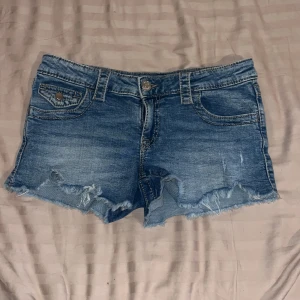Blå low waist jeansshorts från Gina Tricot - Snygga blå jeansshorts från Gina Tricot med fransig och ruffad kant nertill. Klassisk femficksmodell med knappar och dragkedja framtill samt fickor med lock och knapp bak och går även att spänna vid midjan. Perfekta för sommaren och riktigt sköna att bära.