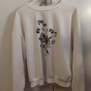 Vit sweatshirt med blommigt tryck - Vit sweatshirt med ett stort blommigt tryck i lila och blå toner på bröstet. Tröjan har rund halsringning och långa ärmar. Mjuk och skön, passar till jeans eller kjol. Kanska stor i passformen.