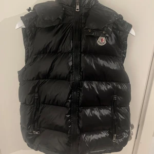 Svart väst från Moncler - Snygg svart dunväst från Moncler med glansig finish och klassisk logga på bröstet. Västen har huva, tvåvägsdragkedja och praktiska fickor med dragkedja. Insidan har ett coolt serietryck. Perfekt för lager-på-lager och streetstyle. Pris kan diskuteras‼️Storlek M (2)