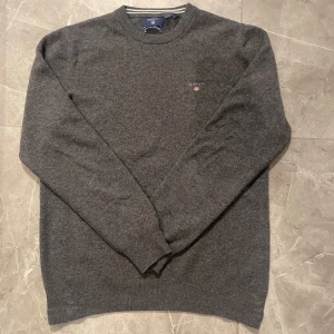 Grå stickad tröja från Gant - Mörkgrå stickad tröja från Gant i superfin lammull. Klassisk rund halsringning med diskret broderad Gant-logga på bröstet. Tröjan har långa ärmar och ribbade muddar för en snygg och enkel stil