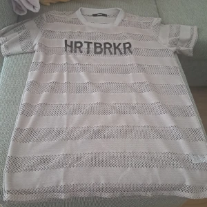 Randig mesh t-shirt HRTBRKR - Säljer en ljusgrå randig t-shirt från Bik Bok med svart HRTBRKR-tryck på bröstet. T-shirten har korta ärmar och är gjord i mesh och bomullsmaterial som ger en luftig känsla. Perfekt för dig som vill sticka ut med en unik och edgy stil. Den är oanvänt. 