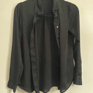Svart långärmad skjorta från H&M - En stilren svart skjorta med långa ärmar och klassisk krage H&M. Skjortan har knäppning framtill och är tillverkad i ett lätt och mjukt material som ger en snygg passform. Perfekt för dig som gillar en clean och enkel look.